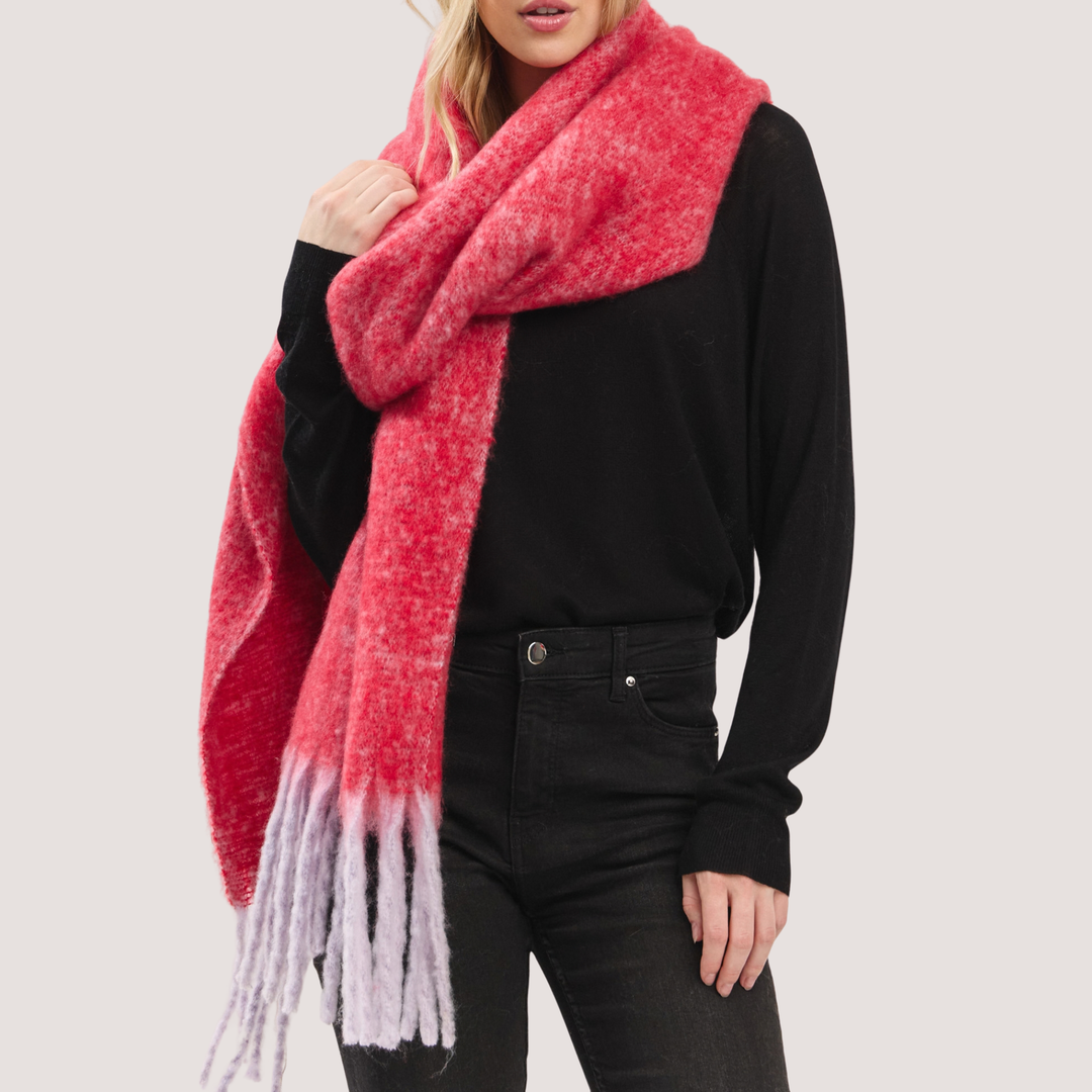 A model wearing a chunky knit scarf with a vibrant red and pink heathered texture and a light lavender-gray ombre fringe, styled over a black long-sleeve top and black jeans against a light gray background.