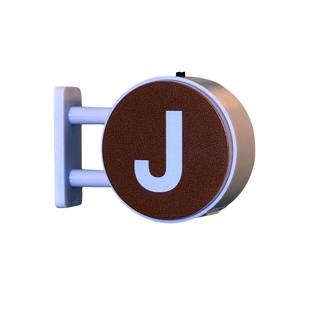 A circular wall-mounted sign with a textured brown face, a large white letter J, and a white mounting bracket, shown on a plain white background.