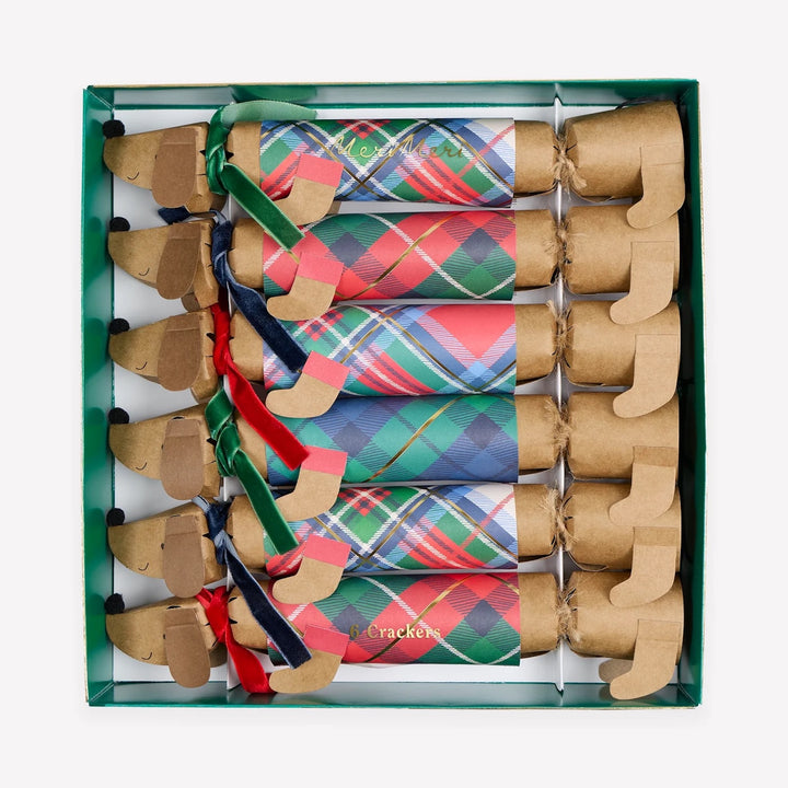 A boxed set of dachshund-shaped paper cracker ornaments with colorful plaid patterns and ribbon accents, arranged inside a green gift box on a light gray background.
