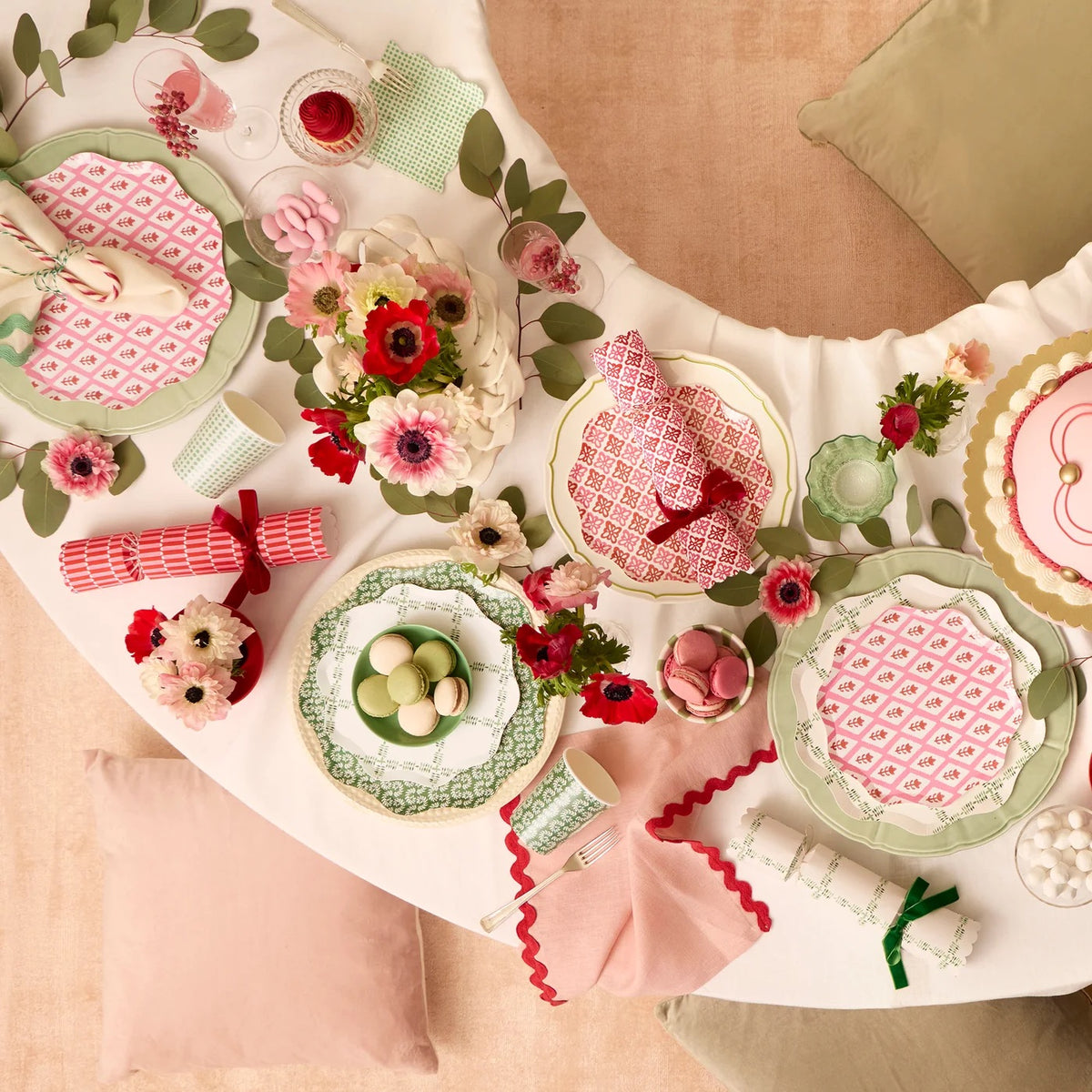 A styled tabletop arrangement with pink patterned napkins, green and white plates, floral centerpieces, red napkins, and small decorative items on a white draped tablecloth, photographed from above.