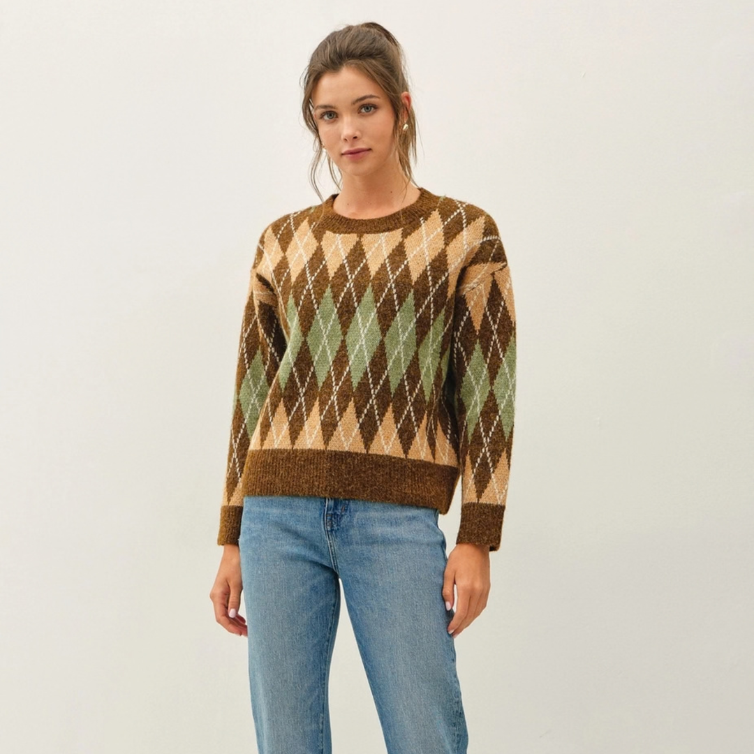 A woman modeling a brown, beige, and green argyle knit sweater with blue jeans, standing against a plain off-white studio background.
