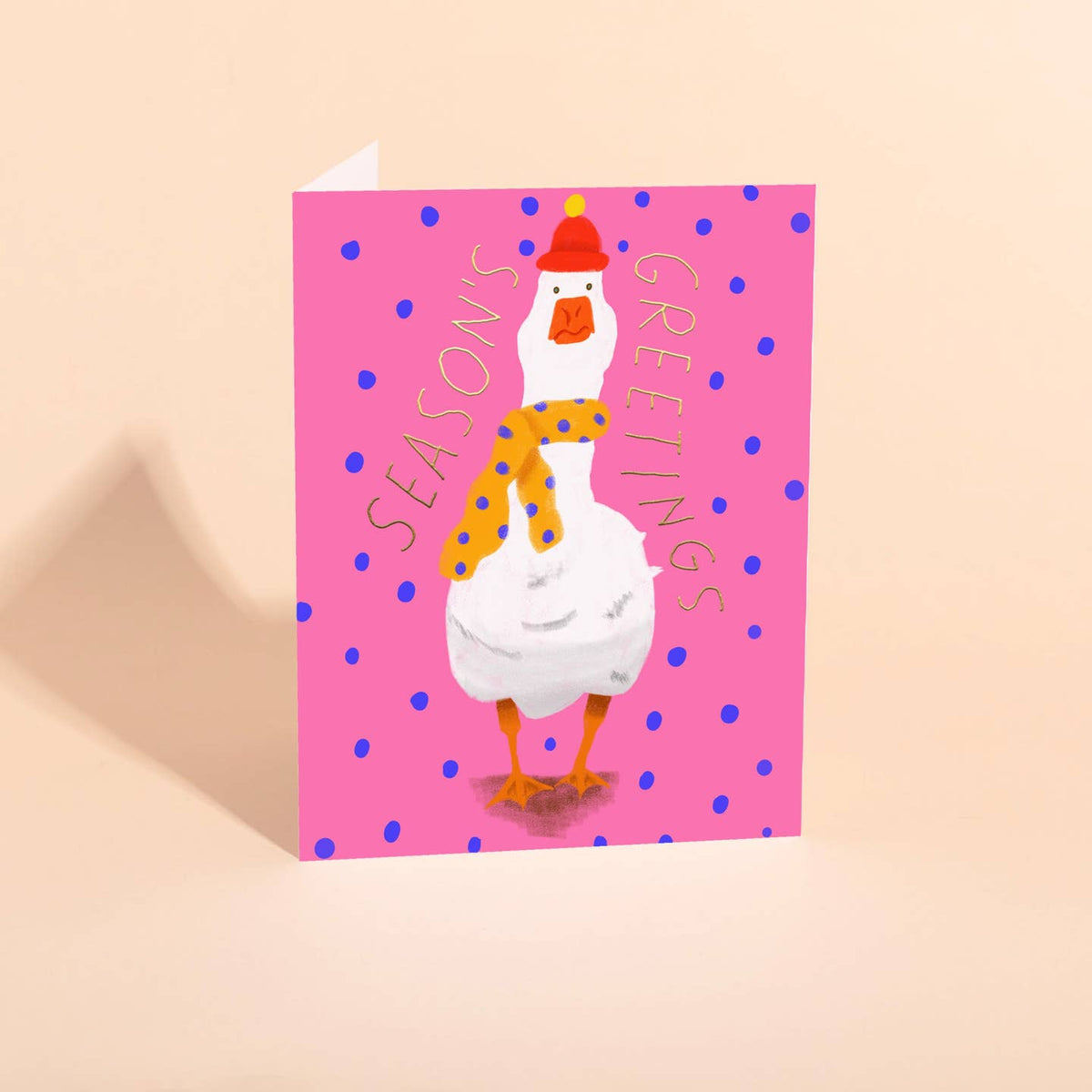 A folded greeting card featuring a white goose wearing a red hat and yellow scarf with blue polka dots, on a pink background with the words 'SEASONS GREETINGS'.