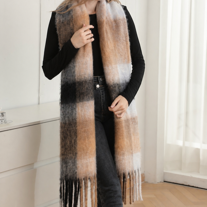 A person wearing a long, fuzzy plaid scarf in beige, brown, black, and white, styled over a black long-sleeve top and dark jeans in a bright indoor setting.