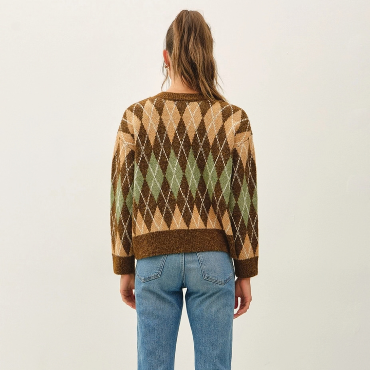 A woman viewed from behind wearing a brown, beige, and green argyle knit sweater with blue jeans, set against a plain off-white studio background.