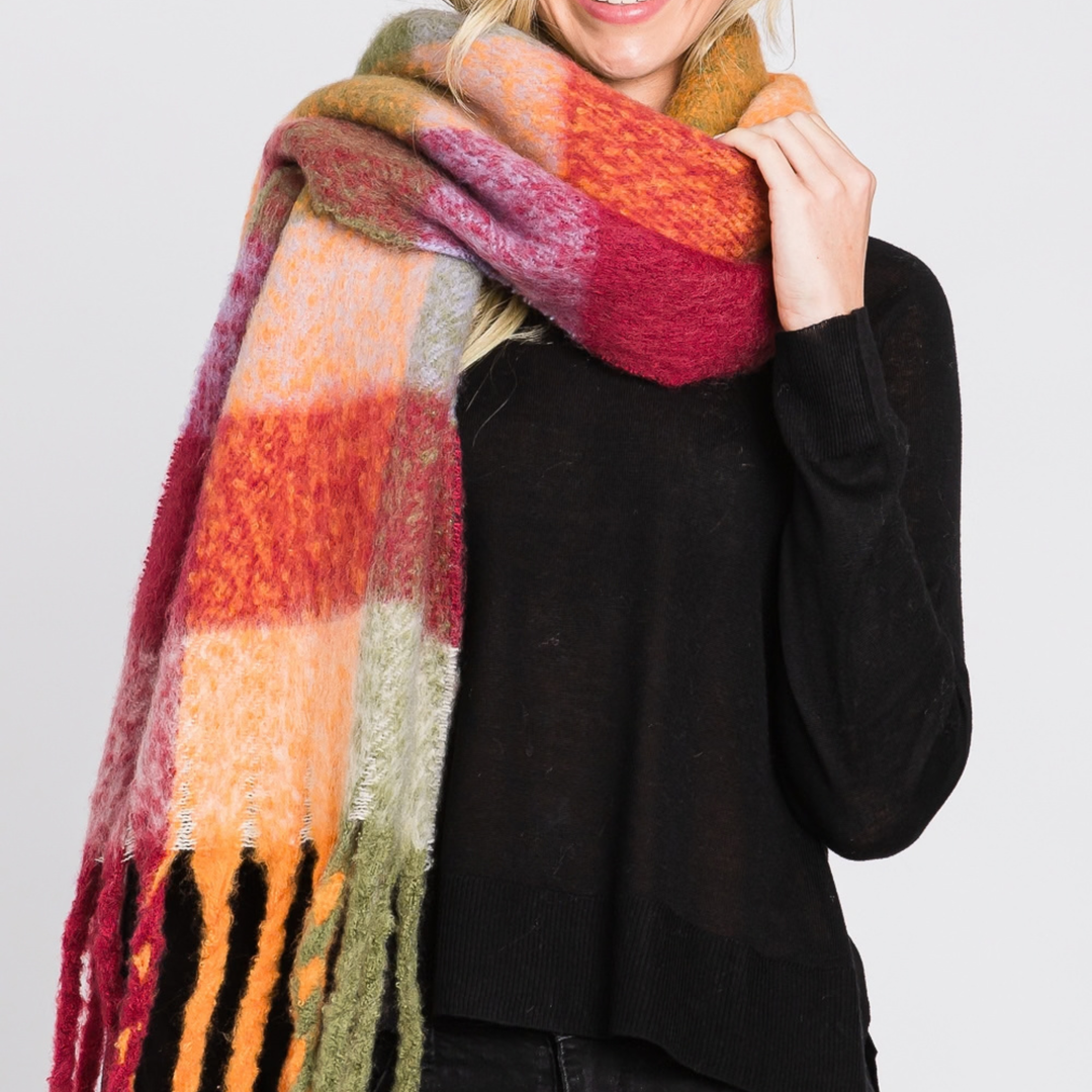 A woman wearing a chunky, fuzzy plaid scarf in warm autumn colors including orange, red, yellow, and green, paired with a black sweater against a light gray background.