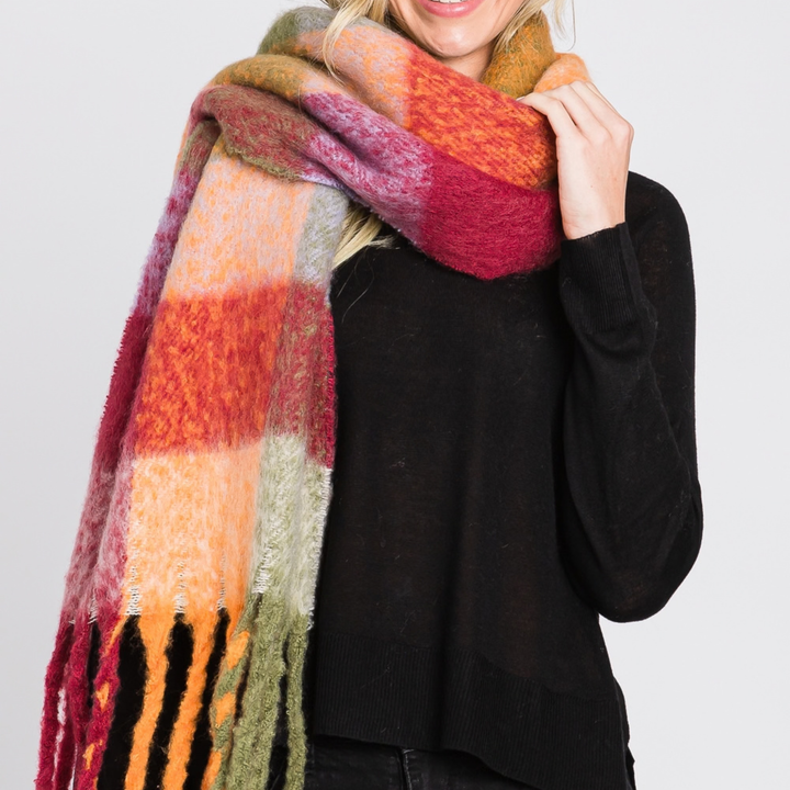 A woman wearing a chunky, fuzzy plaid scarf in warm autumn colors including orange, red, yellow, and green, paired with a black sweater against a light gray background.