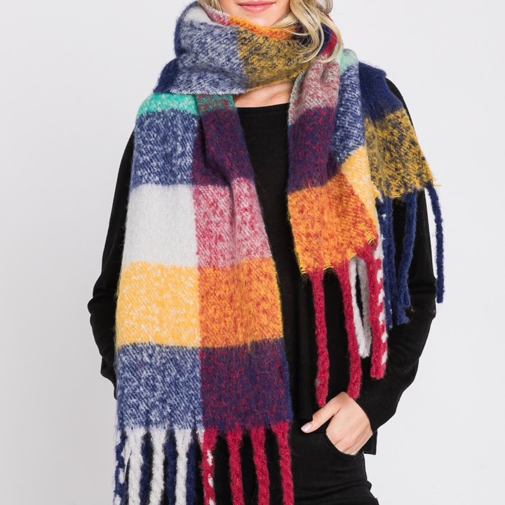 A woman wearing a chunky, oversized plaid scarf with fringe. The scarf features a multicolor checkered pattern of navy blue, mustard yellow, orange, pink, red, white, and teal.