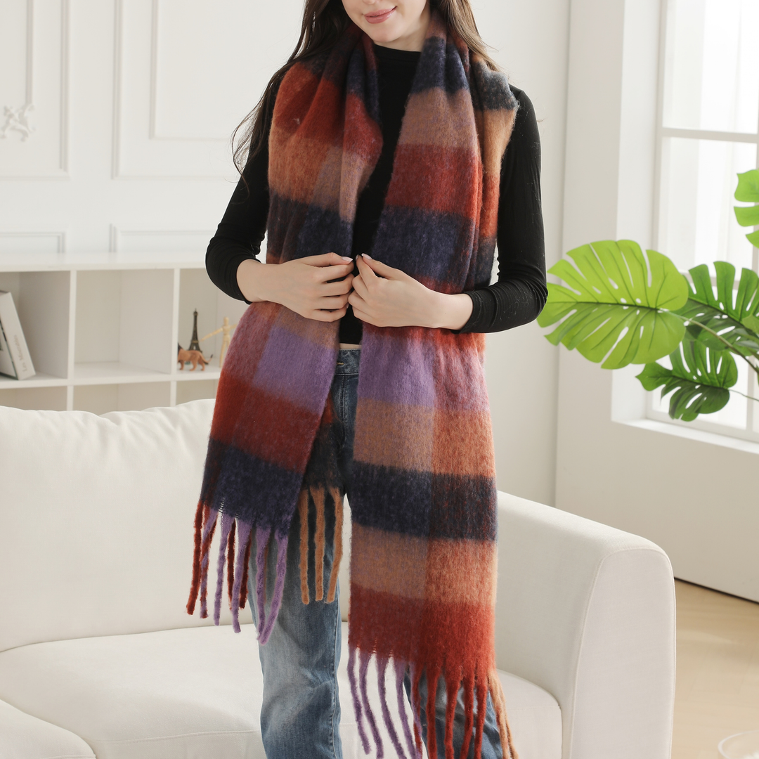 A woman wearing a long, thick plaid scarf in shades of rust red, purple, navy, and beige, paired with a black top and blue jeans, standing in a bright living room next to a white sofa and a green plant.