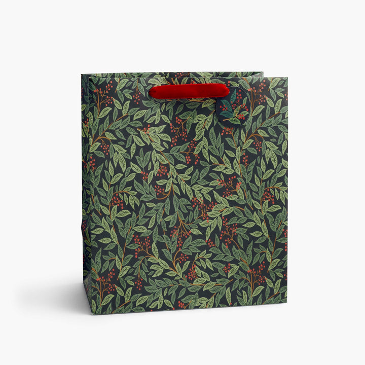 A square gift bag with a dark green and gray botanical pattern of leaves and red berries, featuring a bright red paper handle, shown on a light gray background.
