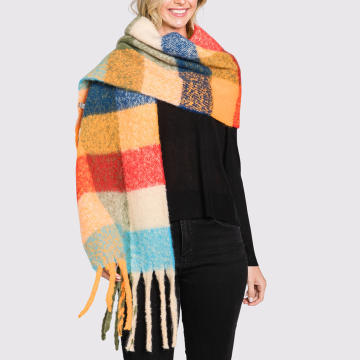 A woman wearing a chunky, oversized plaid scarf in multiple colors including blue, orange, red, yellow, and cream, styled over a black top and black pants against a light gray background.