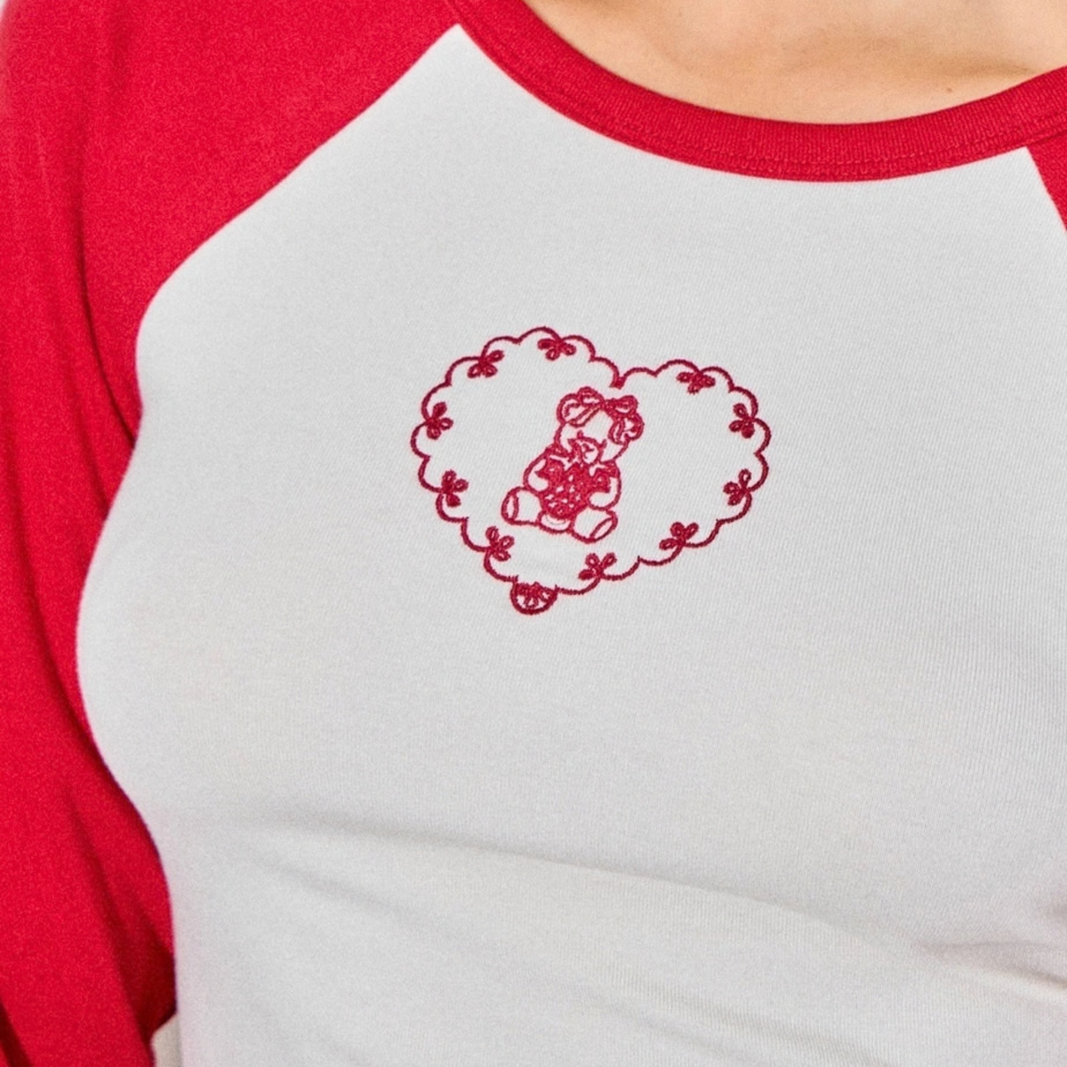 A close-up of a person wearing a white raglan long-sleeve shirt with red sleeves and collar trim, featuring a red embroidered heart with a teddy bear design on the chest.