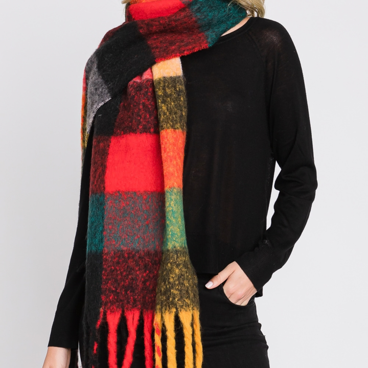 A person wearing a thick, fuzzy plaid scarf in red, green, black, yellow, and orange, paired with a black long-sleeve top against a light gray background.