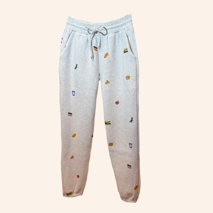 Light heather gray jogger sweatpants with a small multicolor food and drink print, featuring an elastic waistband with a drawstring and side pockets, shown against a solid pale beige background.