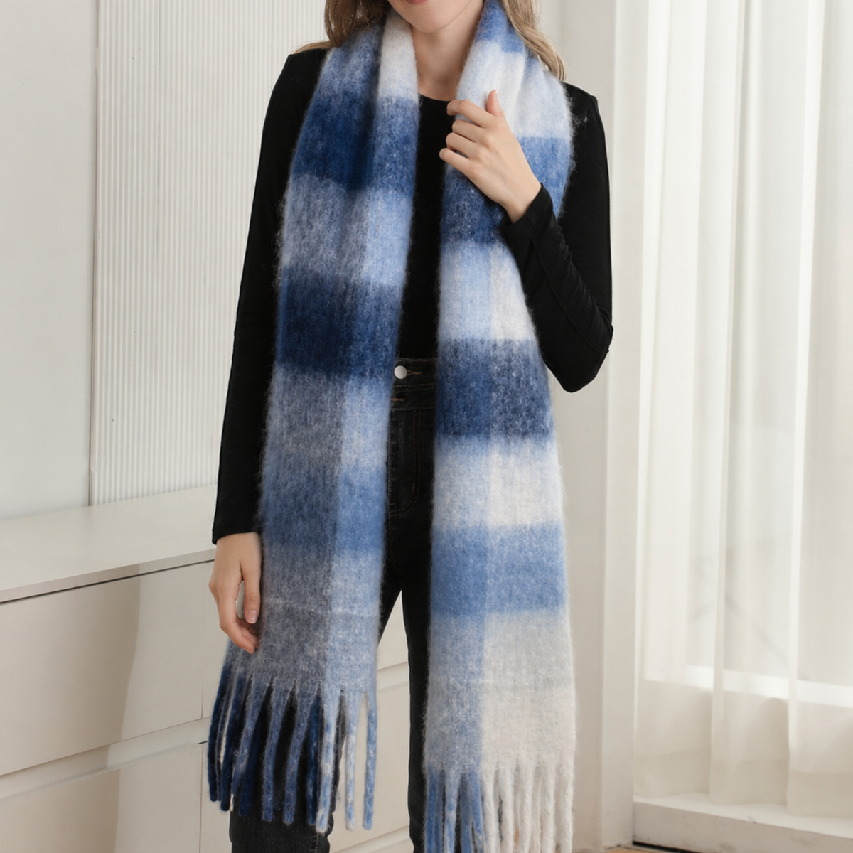 A woman wearing a long, fuzzy plaid scarf in shades of blue, white, and gray, styled over a black top and dark pants.