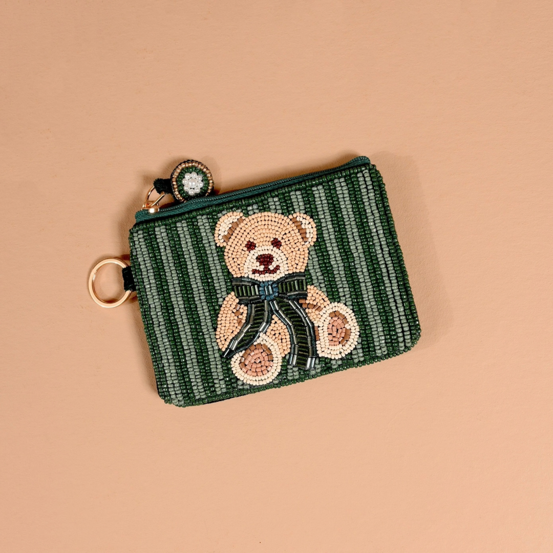 A small zippered coin purse with a beaded teddy bear motif on a dark green and white striped background, featuring a gold-tone key ring and charm, set against a solid peach background.