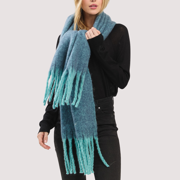 A woman wearing a fuzzy teal-blue wrap poncho with long fringe, styled over a black long-sleeve top and black pants, against a plain light gray background.