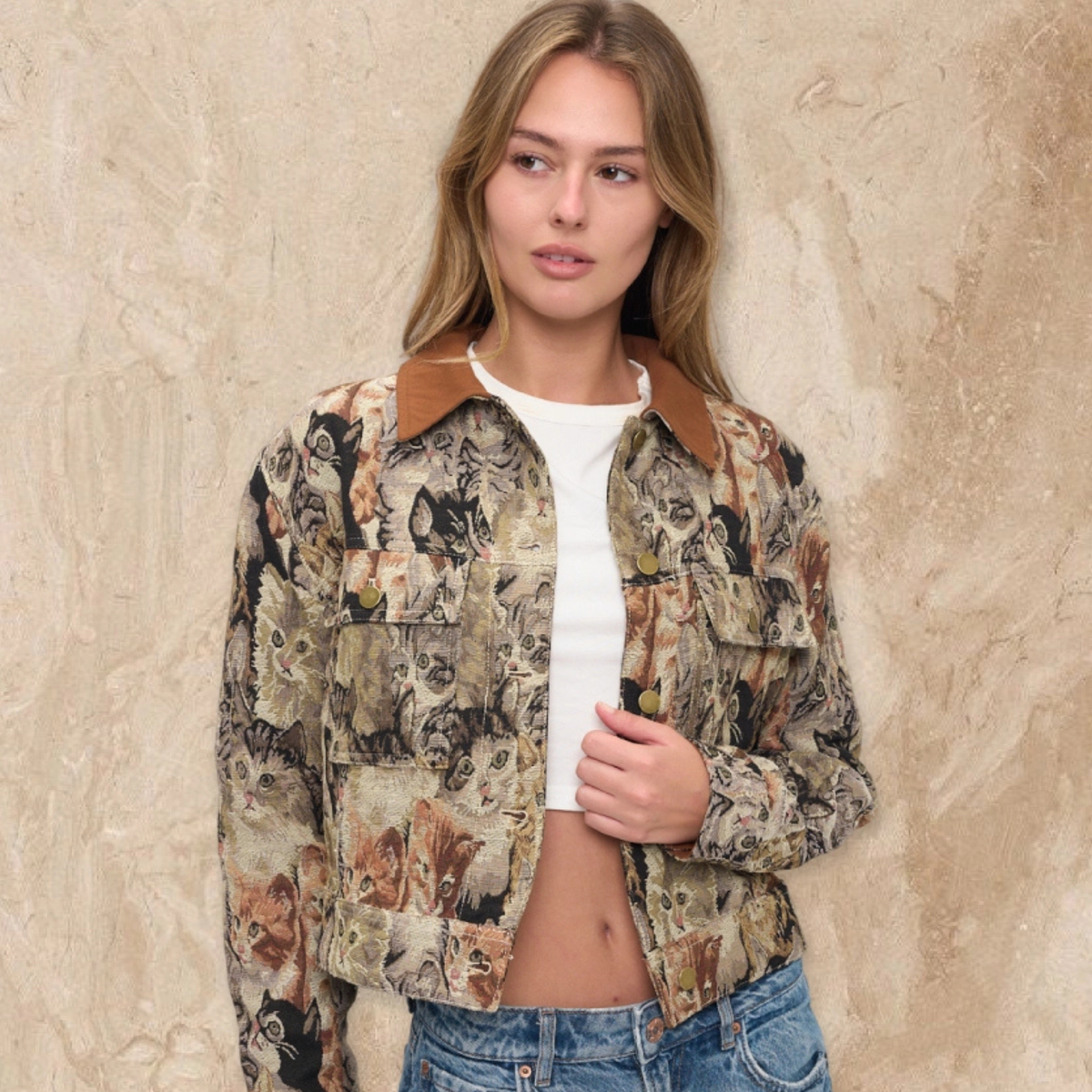 A woman wearing a cropped button-up jacket with an all-over dog print, brown corduroy-style collar, and gold-tone buttons, paired with a white crop top and blue jeans, standing against a textured beige wall.