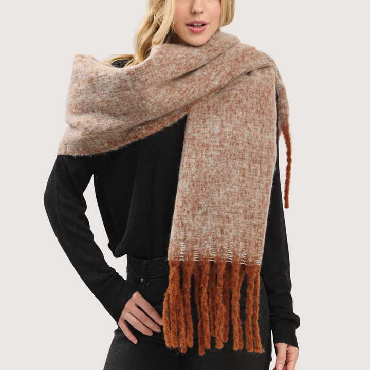 A woman wearing a chunky knit scarf in a heathered beige and brown color, styled over a black long-sleeve top against a light gray background.
