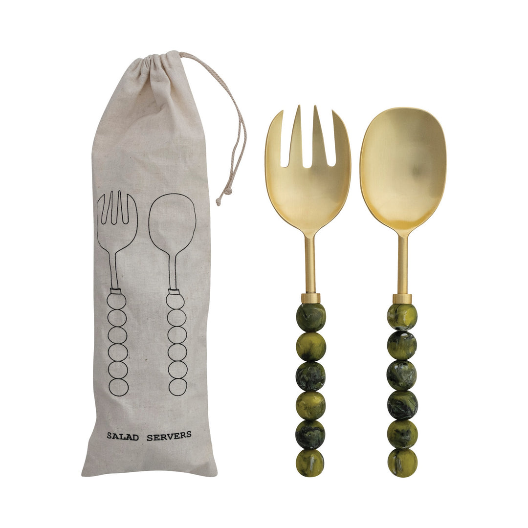 Gold salad servers with green handles next to a drawstring bag labeled &