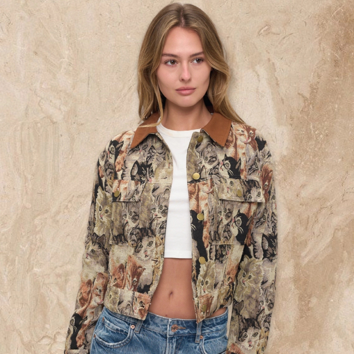A woman wearing a cropped button-up jacket with an all-over dog print, brown collar, and gold buttons, layered over a white crop top and blue jeans, standing against a textured beige wall.