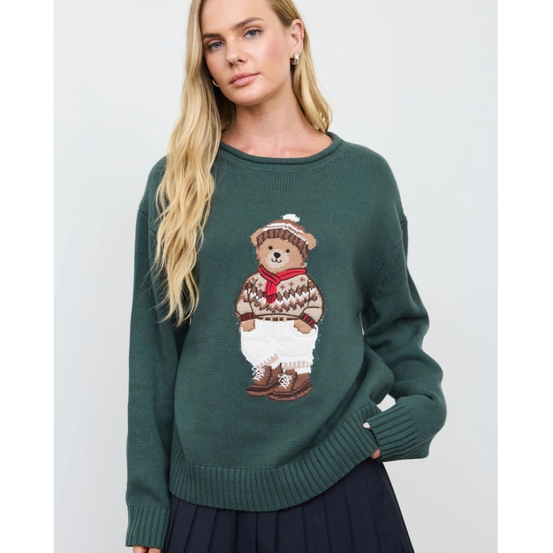 A woman wearing a dark green knit sweater featuring a large embroidered teddy bear in a patterned sweater, white pants, and brown boots on the front, paired with a dark skirt against a light gray background.