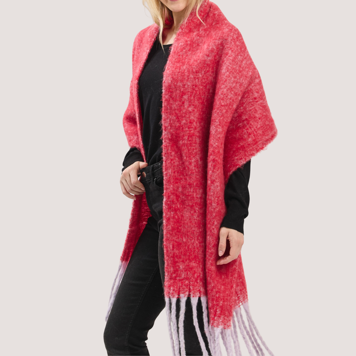 A woman wearing a fuzzy, open-front shawl cape in a heathered red and pink color, styled over a black top and black jeans, against a solid light gray background.