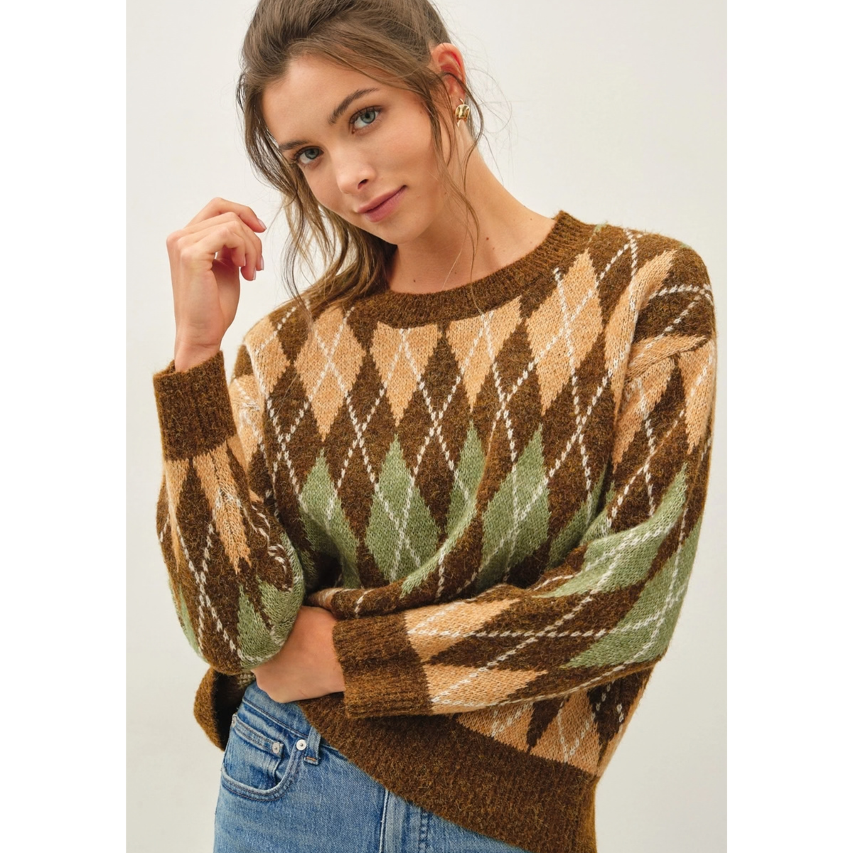 A woman wearing a brown, beige, and green argyle knit sweater with blue jeans, photographed against a plain light background.
