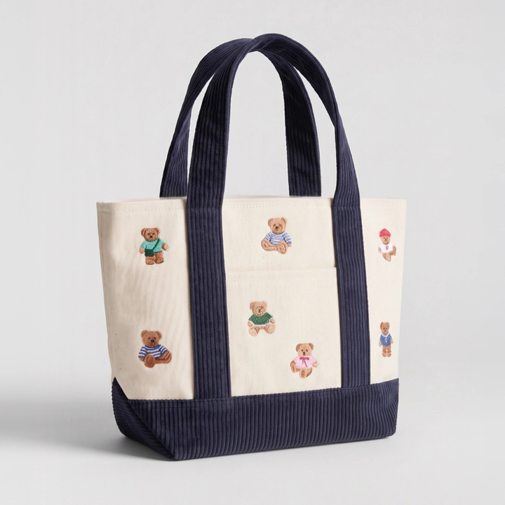 A cream-colored canvas tote bag with wide navy ribbed handles and a matching navy ribbed base, decorated with small embroidered teddy bears in various colorful outfits.