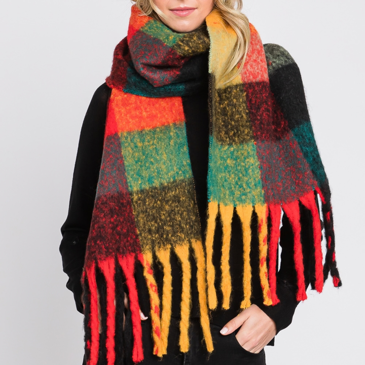 A woman wearing a thick, multicolor plaid scarf with fringe tassels. The scarf features a checkered pattern of red, orange, green, yellow, and black, styled over a black top against a light gray background.