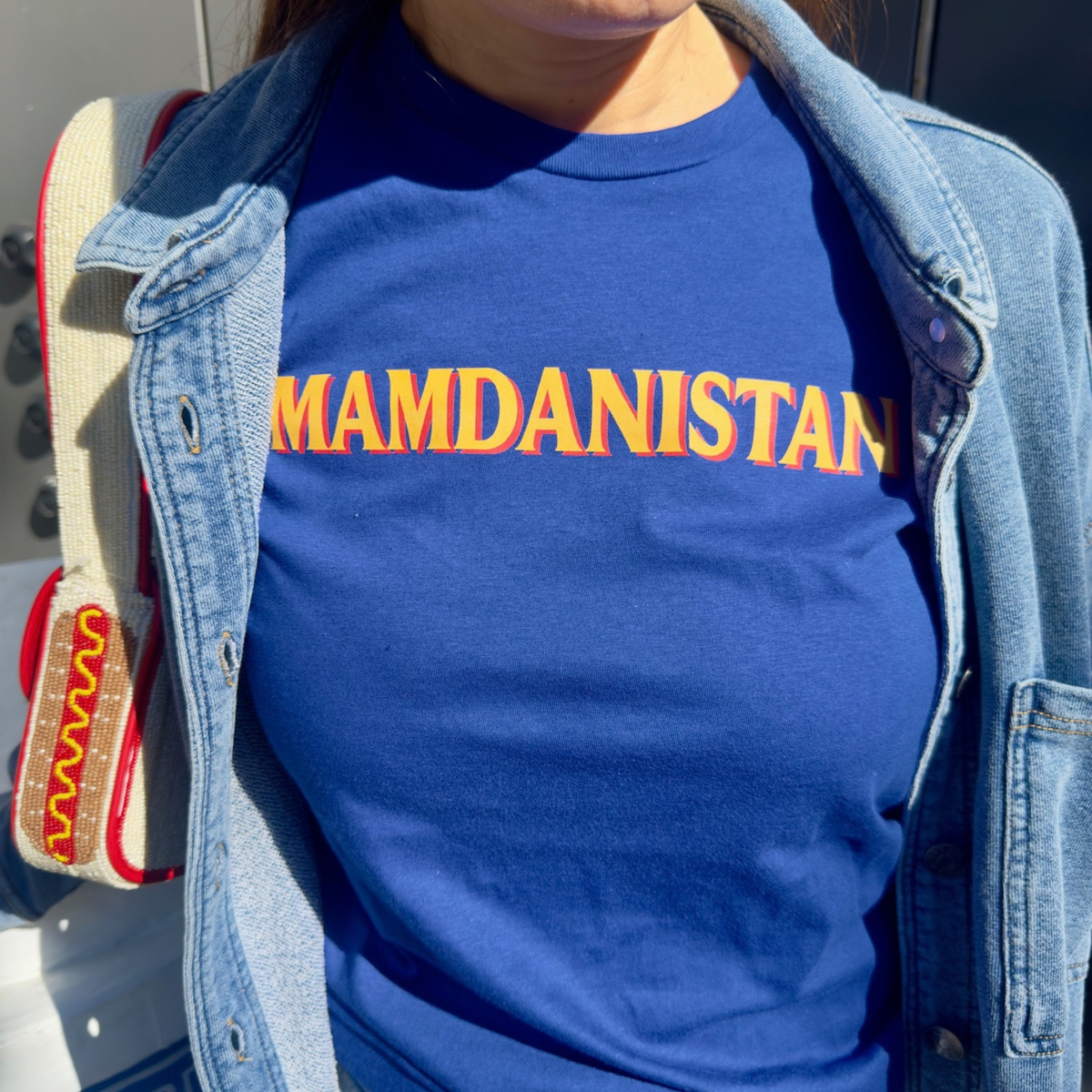 A person wearing a royal blue crewneck t-shirt with 'MAMDANISTAN' printed across the chest in bold orange letters with a yellow outline, styled under an open light-wash denim jacket.