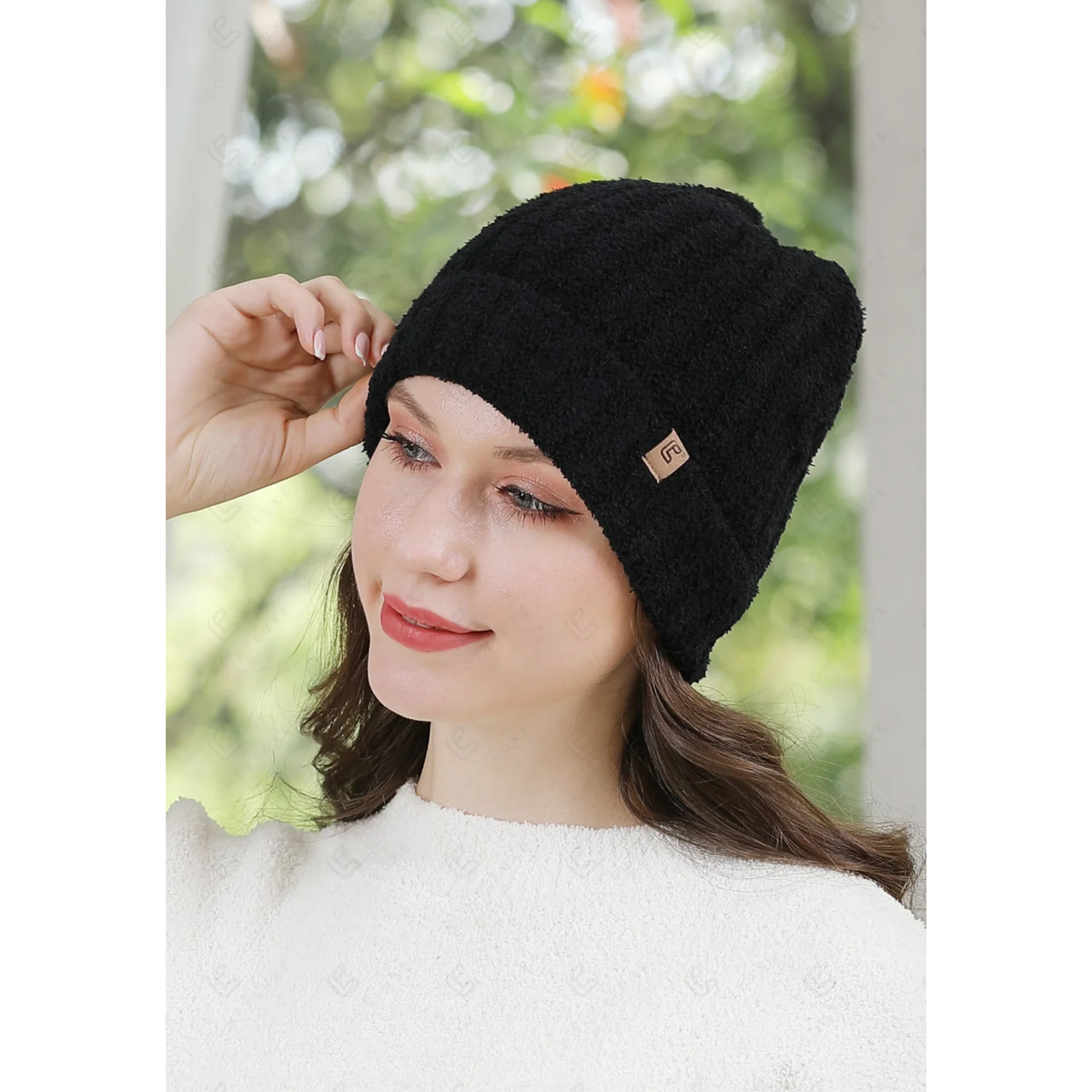 A woman wearing a black fuzzy knit beanie hat with a small tan brand patch on the side, posed against a soft-focus outdoor background.