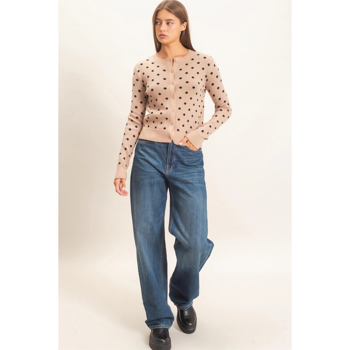 A woman modeling a beige zip-up cardigan with black polka dots, paired with wide-leg blue jeans and black shoes, against a plain light beige studio background.