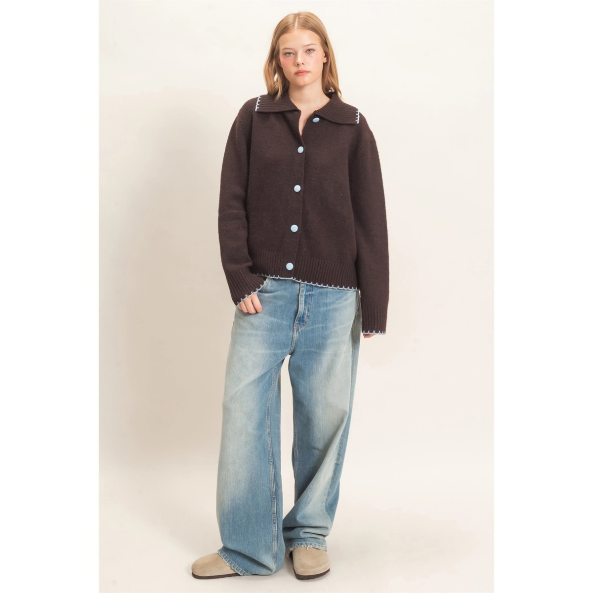 A woman modeling a dark brown knit cardigan with light blue buttons and decorative trim, paired with wide-leg light blue jeans and beige shoes, against a plain beige studio background.
