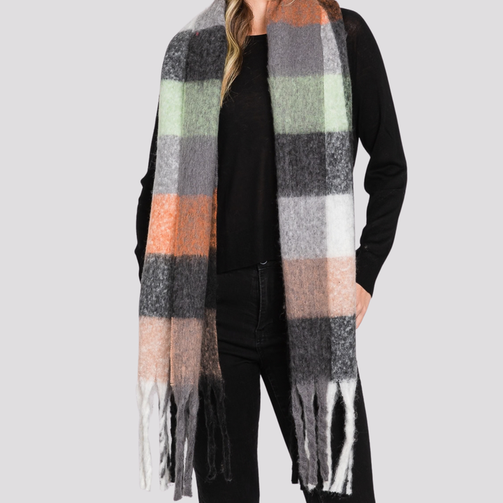 A woman wearing a long, fuzzy plaid scarf with fringe ends. The scarf features large blocks of gray, white, black, peach, orange, and pale green, styled over a black top and black pants against a solid light gray background.