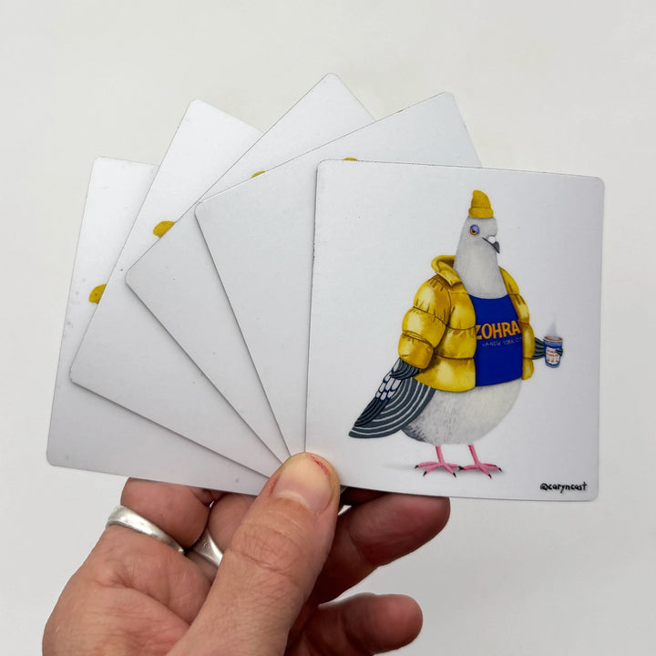 A hand holds a fanned-out set of square cards against a plain light background. The front card shows an illustrated pigeon wearing a yellow puffer jacket and blue shirt with the word "CHOSEN" on it.