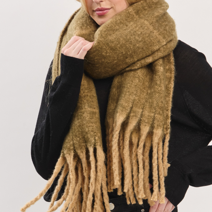 A close-up of a person wearing a thick, fuzzy mustard-brown fringed scarf over a black top, against a plain light gray background.