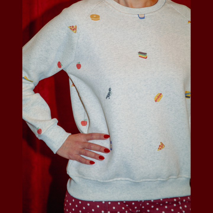 A person wearing a light heather gray sweatshirt with small colorful food-themed patches, posed against a dark red curtain background.