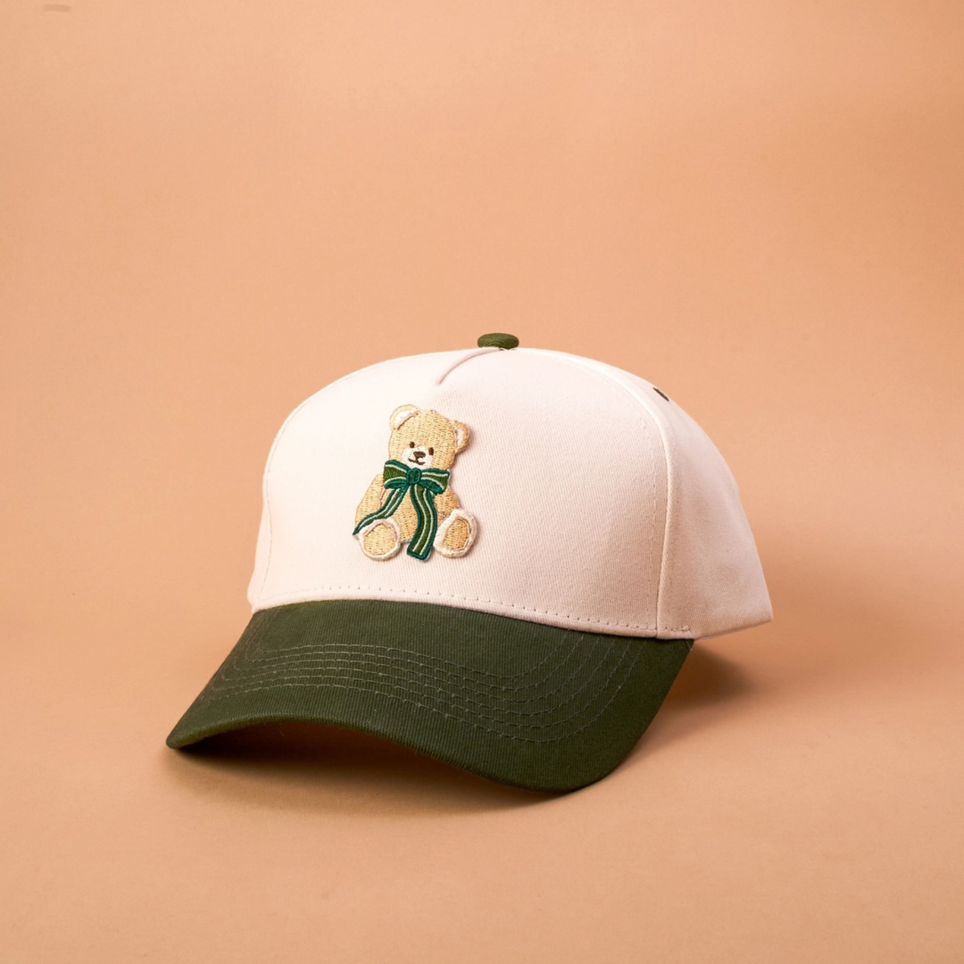A baseball cap with an off-white crown, a dark green curved brim, and a small embroidered teddy bear wearing a green scarf on the front, set against a solid peach background.