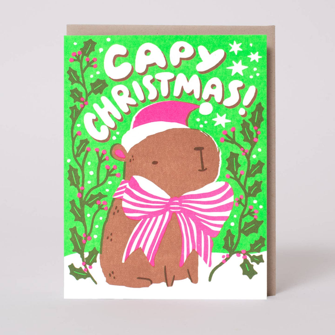 A square Christmas greeting card featuring a cartoon capybara wearing a pink Santa hat and matching pink striped bow, with the words “Capybara Christmas!” on a bright green background with holly and stars. The card is shown standing upright with a kraft envelope behind it on a light gray background.