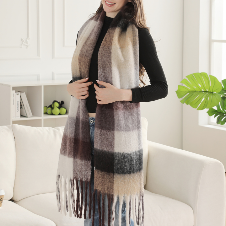 A woman wearing a long, soft plaid scarf in neutral tones of beige, brown, gray, and black, styled over a black top and blue jeans while standing in a bright living room next to a white sofa.