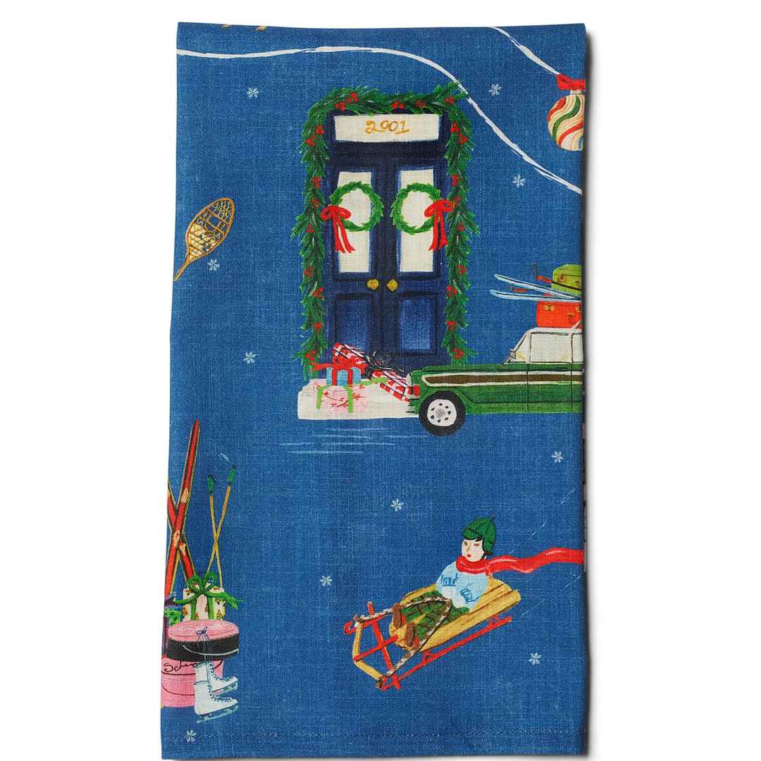 A blue fabric tea towel featuring a festive Christmas-themed illustration with a decorated door, a green car, a snowboard, skis, and a person sledding.