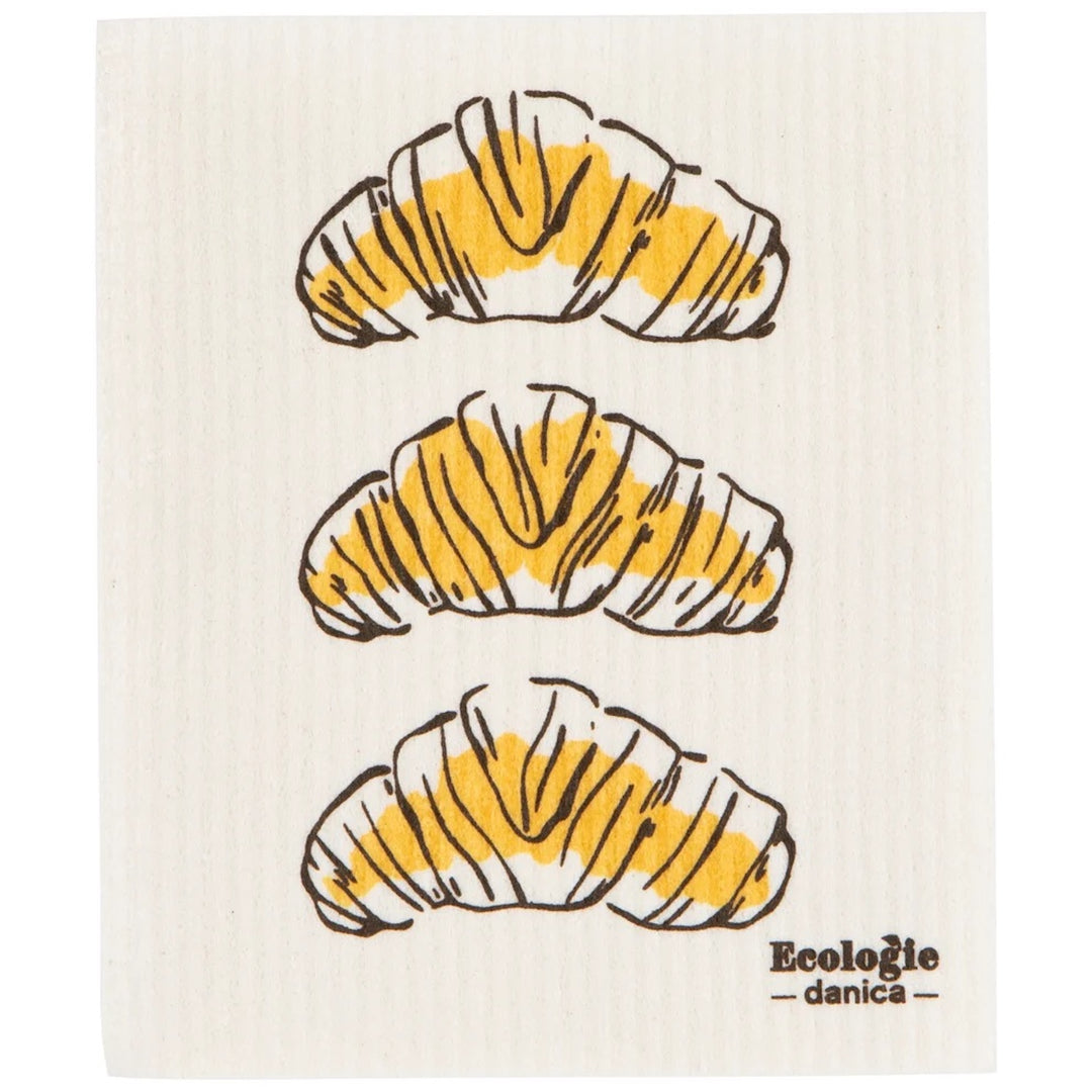 A square white dishcloth with a textured, ribbed surface, printed with three croissant illustrations in yellow and brown, and the brand name &